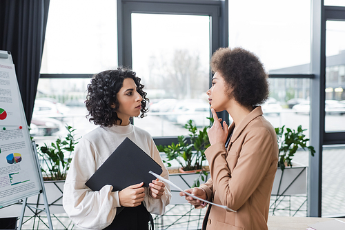Pensive interracial businesswomen holding newspaper and paper folder in office