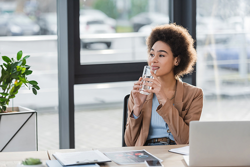 Smiling african american businesswoman holding glass of water near devices and papers in office