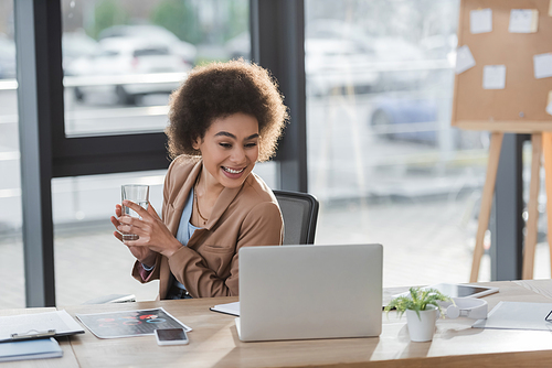 Cheerful african american businesswoman holding glass of water near laptop and documents in office