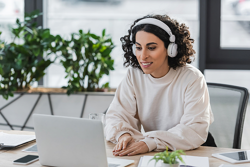 Smiling muslim businesswoman in headphones looking at laptop in office