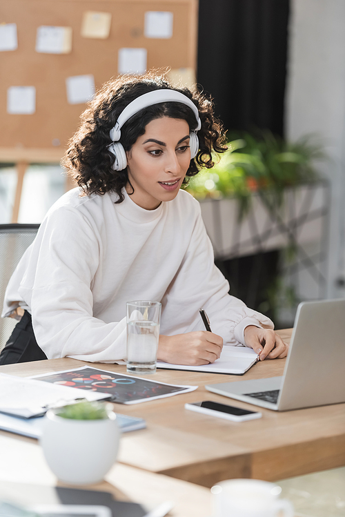 Muslim businesswoman in headphones writing on notebook near devices in office
