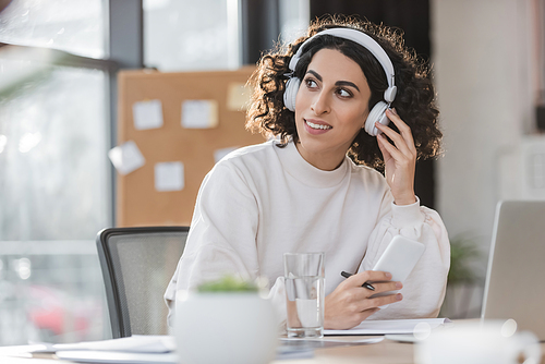 Cheerful muslim businesswoman in headphones holding pen and cellphone in office