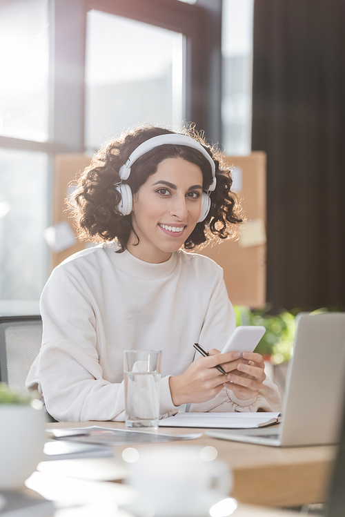 Smiling arabian businesswoman in headphones holding cellphone and looking at camera in office