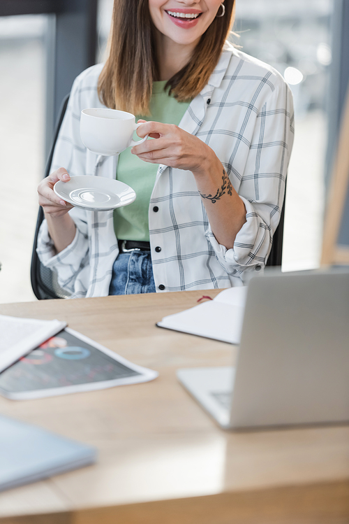 Cropped view of smiling businesswoman holding cup of coffee near blurred laptop in office