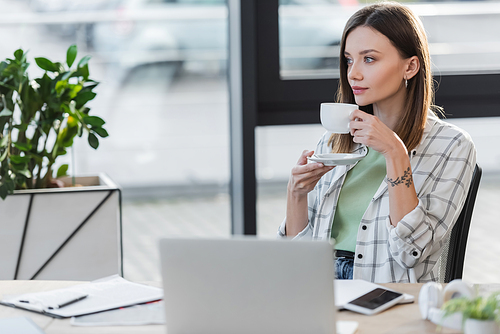 Young businesswoman holding cup near blurred devices in office