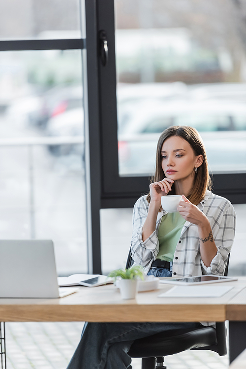 Pensive businesswoman holding cup near blurred laptop in office