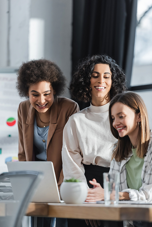 Happy multicultural businesswomen looking at blurred laptop in office