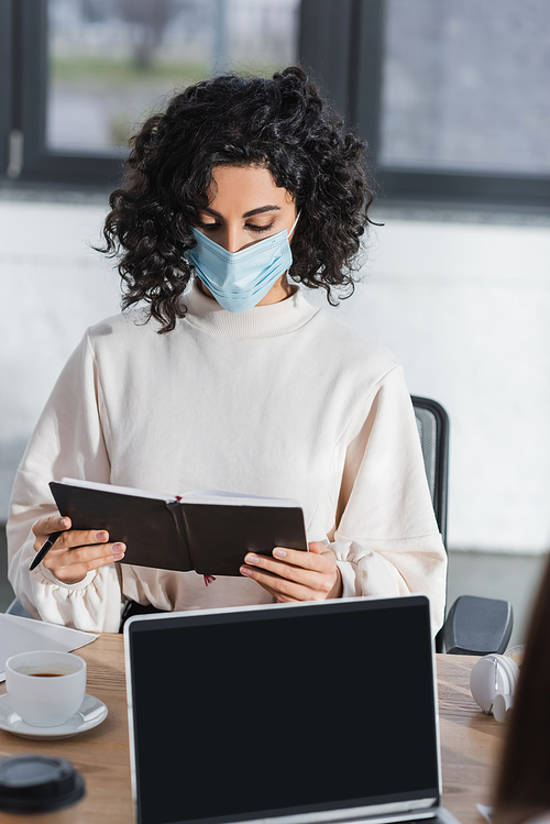 Arabian businesswoman in medical mask holding notebook near laptop and coffee in office
