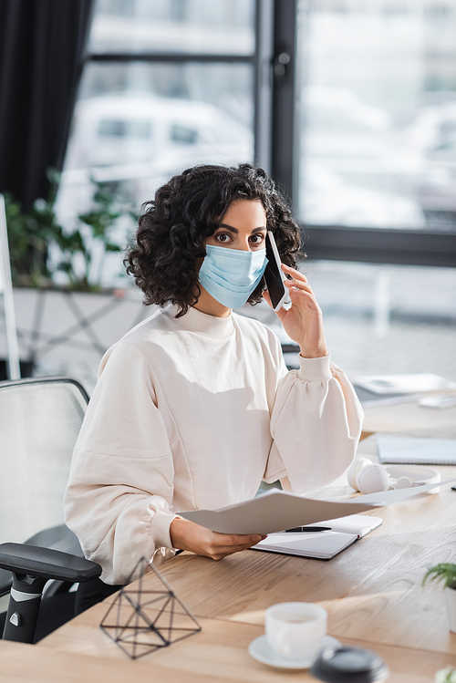 Muslim businesswoman in medical mask talking on cellphone and holding documents in office