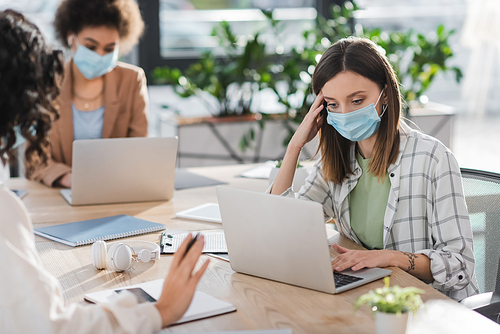 Businesswoman in medical mask using laptop near blurred interracial colleagues in office