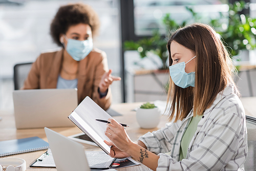 Businesswoman in medical mask holding notebook near blurred laptop and african american colleague in office