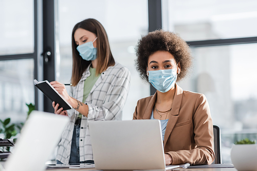 African american businesswoman in medical mask looking at camera near laptop and colleague in office