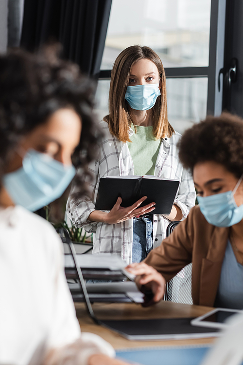 Businesswoman in medical mask holding medical mask near blurred interracial colleagues working in office