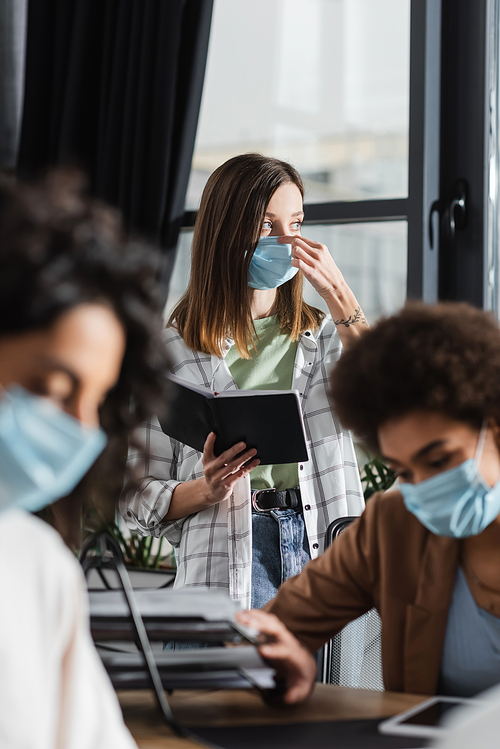 Young businesswoman holding notebook and wearing medical mask near blurred multiethnic colleagues in office