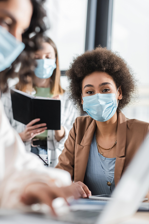 African american businesswoman in medical mask looking at camera near blurred interracial colleagues in office