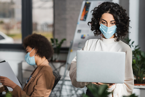 Muslim businesswoman in medical mask using laptop near blurred african american colleague in office