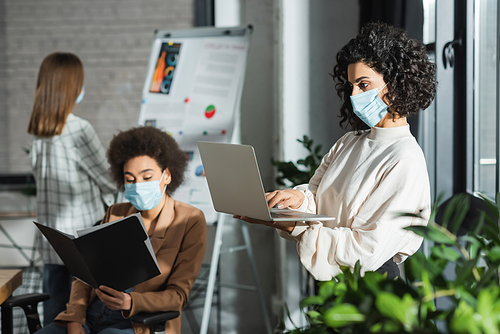 Arabian businesswoman in medical mask using laptop near plant and interracial colleagues in office