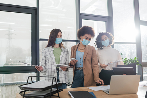 Multiethnic businesswomen in medical masks holding devices and working with papers in office