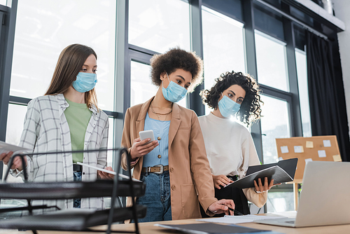 Interracial businesswomen in protective masks holding gadgets and working with papers in office