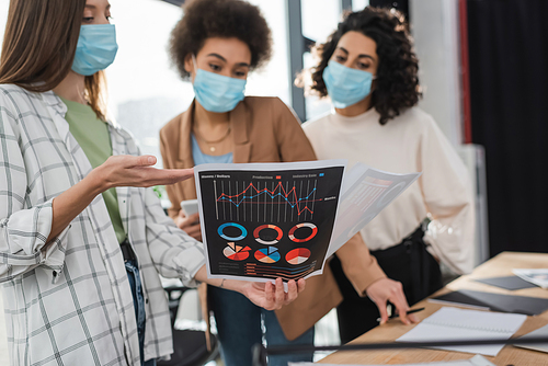 Businesswoman in medical mask pointing at paper with charts near blurred multiethnic colleagues in office
