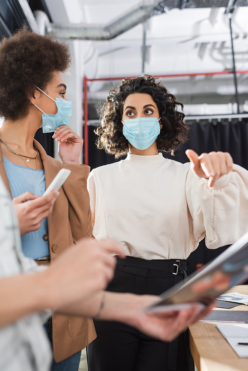 Arabian businesswoman in medical mask pointing at blurred paper near interracial colleagues in office