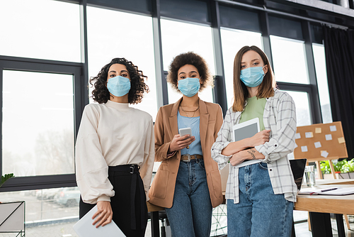 Interracial businesswomen in medical masks holding devices and papers in office