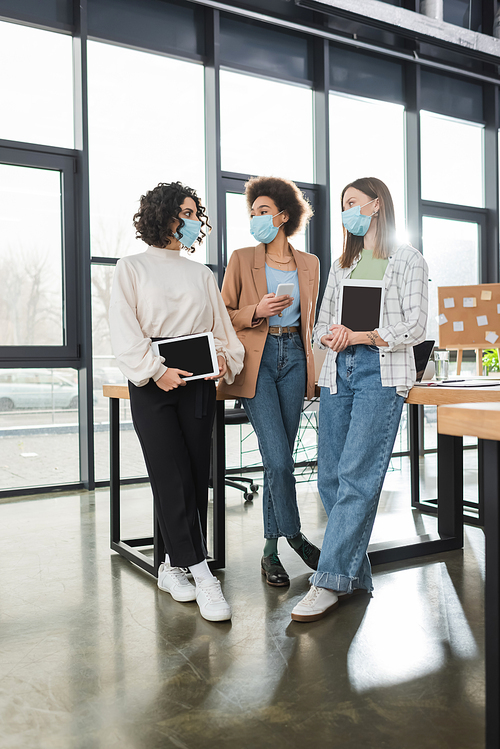 Multiethnic businesswomen in medical masks holding devices while talking in office