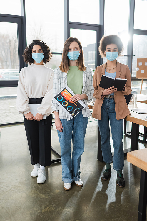 Multicultural businesswomen in protective masks holding digital tablet, paper and notebook in office