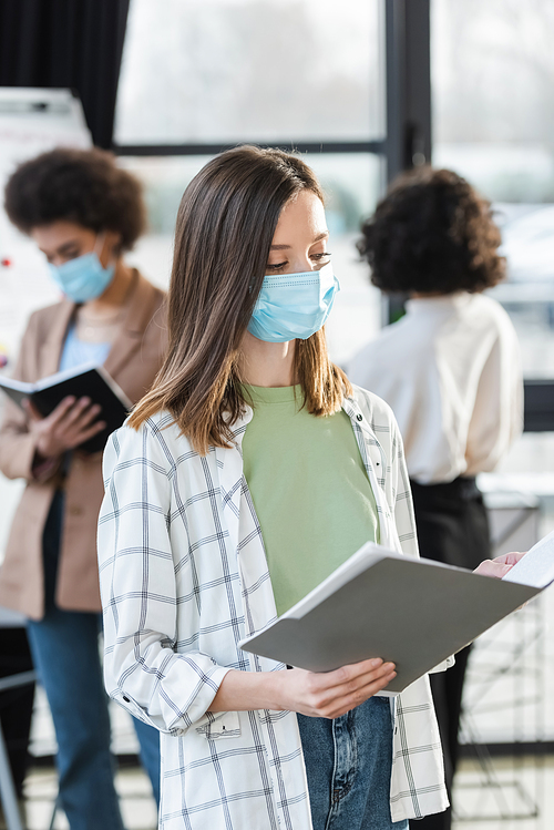 Businesswoman in medical mask holding blurred paper folder near blurred interracial colleagues in office