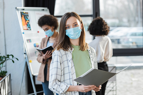Young businesswoman in medical mask holding paper folder near blurred multiethnic colleagues