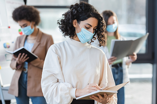 Arabian businesswoman in medical mask writing on notebook near blurred colleagues in office