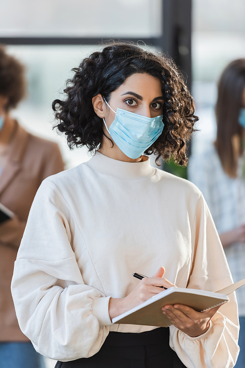 Arabian businesswoman in protective mask holding pen and notebook while looking at camera in office