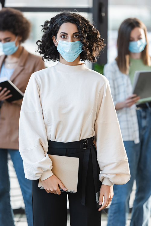 Muslim businesswoman in medical mask holding notebook and looking at camera in office