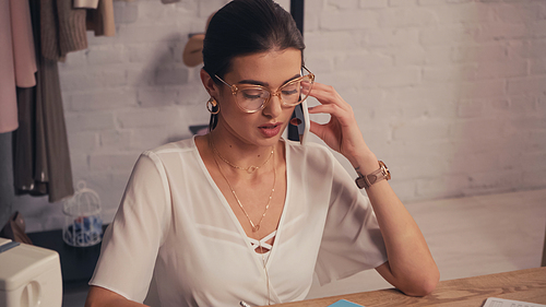 Seamstress in eyeglasses talking on smartphone in atelier