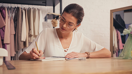 Brunette designer in eyeglasses writing on notebook in atelier