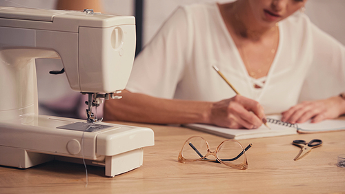 Cropped view of sewing machine near eyeglasses and blurred seamstress in atelier