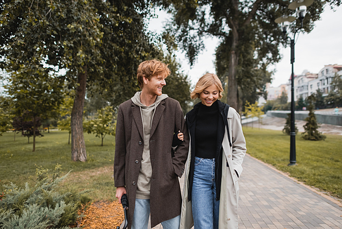 young and positive man in coat walking with blonde girlfriend while holding umbrella in autumnal park