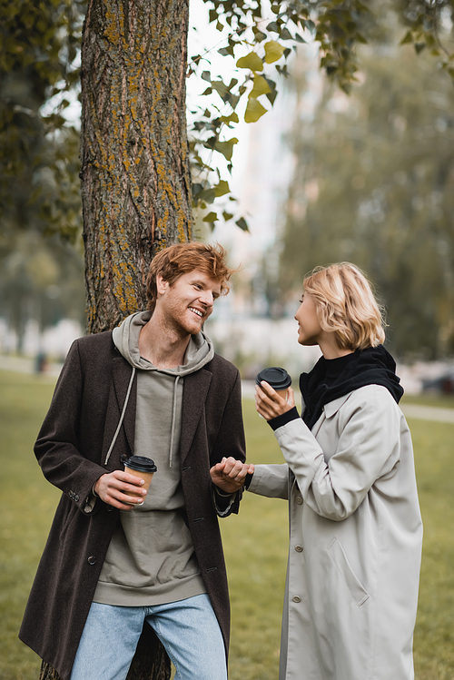 positive man in coat holding paper cup and looking at pleased girlfriend neat tree trunk in autumnal park