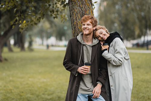 happy blonde woman holding paper cup and leaning on shoulder of smiling boyfriend near tree trunk