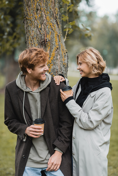 happy blonde woman holding paper cup and laughing with boyfriend near tree trunk