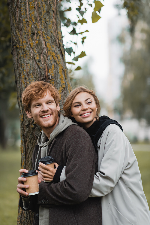 happy blonde woman holding paper cup and hugging with boyfriend near tree trunk