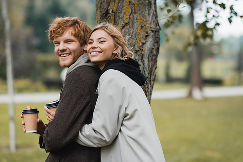 cheerful blonde woman holding paper cup and hugging with boyfriend near tree trunk