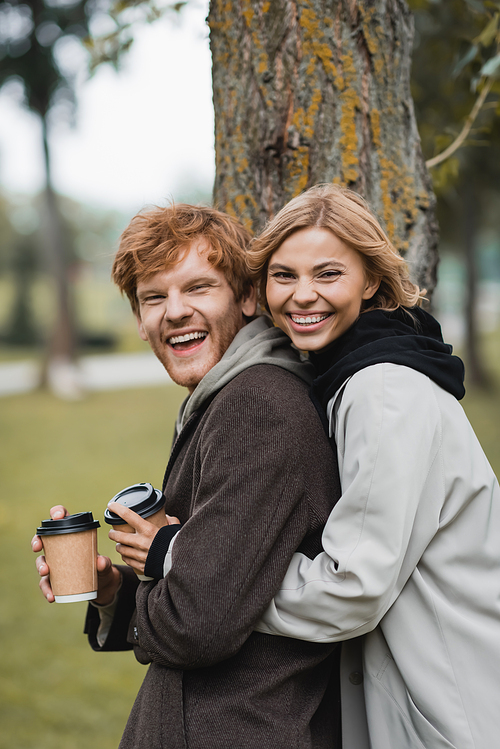 cheerful blonde woman holding paper cup and hugging with positive boyfriend near tree trunk