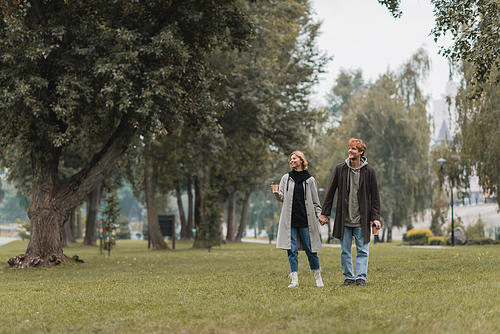 full length of happy redhead man and cheerful woman holding coffee to go while walking in park