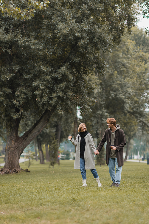 full length of positive redhead man and cheerful woman holding coffee to go while walking in park