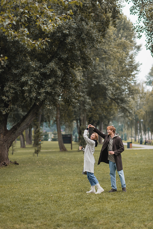 full length of happy redhead man and cheerful woman holding coffee to go while dancing in park