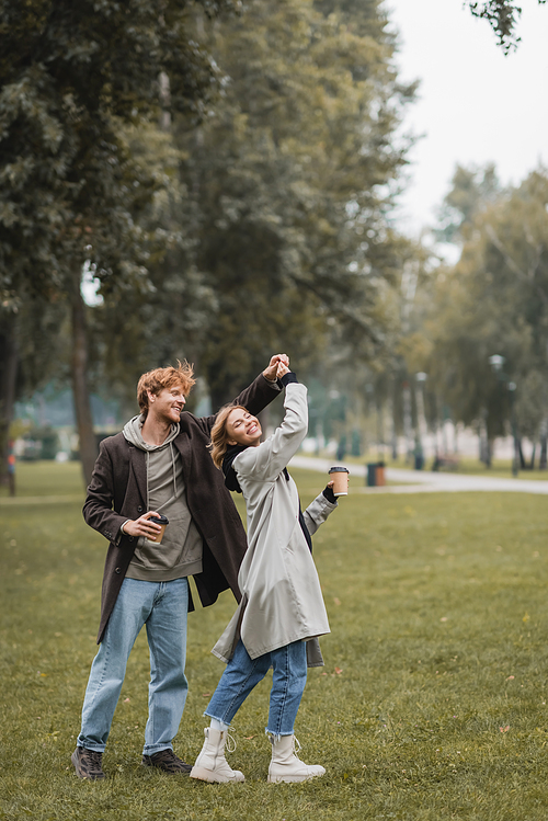 full length of happy redhead man and cheerful woman holding coffee to go while twirling during dance in park