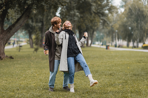 full length of happy redhead man and cheerful woman holding coffee to go while laughing in park