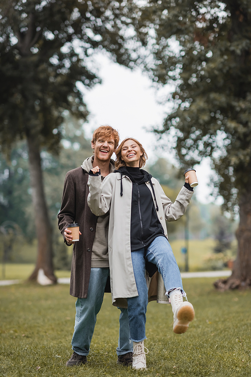 full length of happy redhead man and pleased young woman holding coffee to go while walking in park