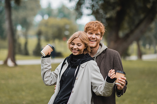 happy redhead man and pleased young woman holding coffee to go while walking in park